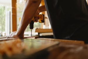 Crop anonymous male carpenter making hole on wooden plank with drill during work in studio on sunny day — Affordable MDF Bull
